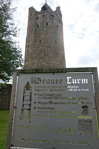 Hoher, steinerner "Grauer Turm" mit Infotafel im Vordergrund, die Merkmale und Öffnungszeiten des mittelalterlichen Turms zeigt.