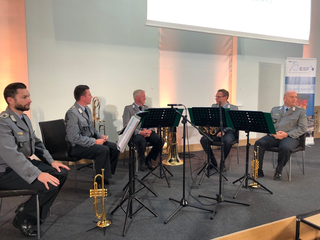 Fünf Männer in Uniformen mit Blechblasinstrumenten sitzen auf einer Bühne, möglicherweise eine Militärkapelle.