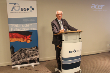 Mann spricht auf GSP-Veranstaltung, mit Banner der deutschen Flagge, Reichstag und 70-jährigem Jubiläumslogo der Organisation.