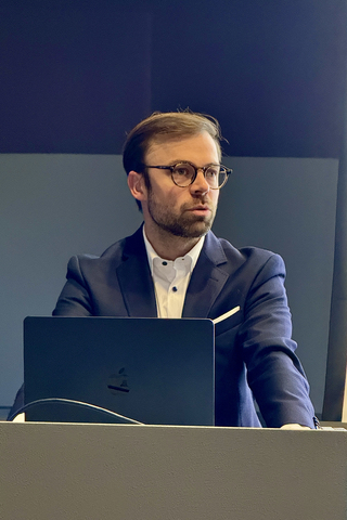 Mann in dunklem Anzug und Brille, mit einem Laptop vor sich, blickt während einer Präsentation nach rechts.