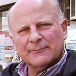 Jürgen Kahl, Düsseldorf Nahaufnahmeporträt eines älteren, glatzköpfigen Mannes mit leichtem Lächeln, der eine dunkle Jacke über einem gestreiften Hemd trägt.