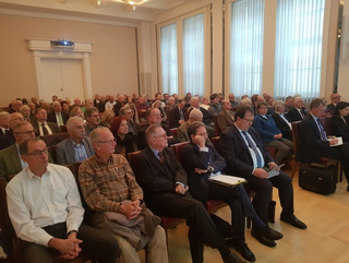 Eine große Gruppe von Erwachsenen sitzt in Reihen in einem Konferenzsaal und hört einer Präsentation zu.