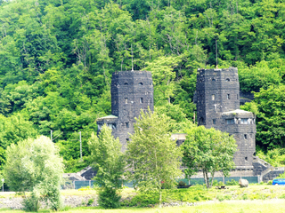Zwei dunkle Steintürme der Brücke von Remagen, umgeben von dichtem grünem Wald und Gras unter hellem Himmel.