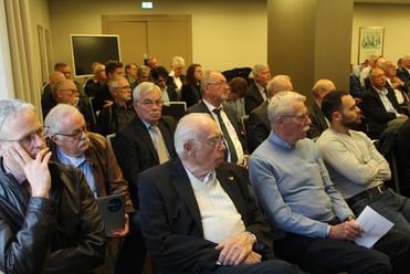 Eine Gruppe von überwiegend Männern verschiedenen Alters sitzt aufmerksam in Reihen bei einer Konferenz oder Präsentation.