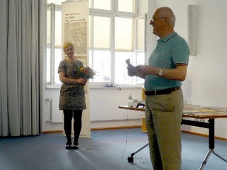 Eine Frau mit Blumenstrauß lächelt einen älteren Mann an, der einen Gegenstand hält, bei einer Veranstaltung in einem Raum mit Fenstern.