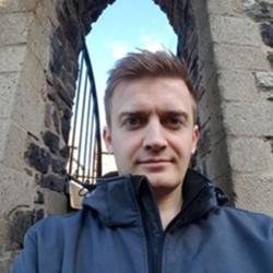 Ein Mann in einer dunklen Jacke macht ein Selfie vor einem alten Steinbogen, durch den blauer Himmel zu sehen ist.