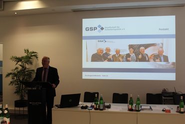 Ein Mann spricht an einem Pult vor einer Projektionsfläche, die das GSP-Logo und ein Gruppenfoto zeigt.