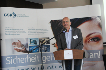 Mann am Rednerpult vor einem Banner der Gesellschaft für Sicherheitspolitik (GSP), das über die Sicherheitslage informiert.