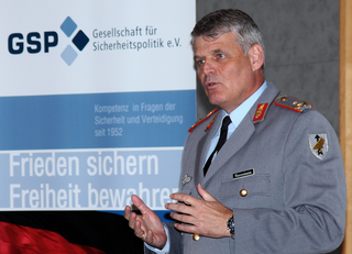 Oberst Hannemann spricht in Uniform vor einem GSP-Banner über Sicherheitspolitik.