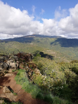 Ein Wanderweg entlang eines felsigen Randes mit Blick auf eine weite Landschaft bewaldeter Berge unter teilweisem Himmel.