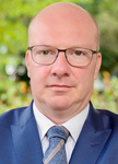 Porträt eines glatzköpfigen Mannes mit Brille, blauem Anzug und Krawatte, blickt direkt in die Kamera.