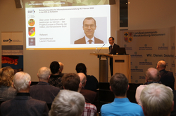 Generalkonsul Toulouse spricht an einem Pult vor Publikum, mit einer Sicherheitspolitik-Präsentation im Hintergrund.