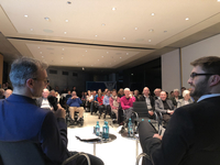 Zwei Redner sprechen vor einem gemischten Publikum, teils in Rollstühlen, in einem modernen Konferenzraum. Einer hält ein Mikrofon.