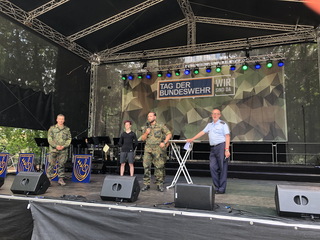 Vier Personen, davon drei in Uniform, stehen auf einer Bühne mit dem Banner 'Tag der Bundeswehr Wir sind da' im Hintergrund.