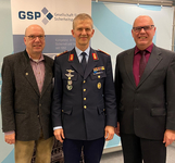 Drei lächelnde Männer: Einer in Militäruniform, zwei in Zivil, vor einem GSP-Banner.