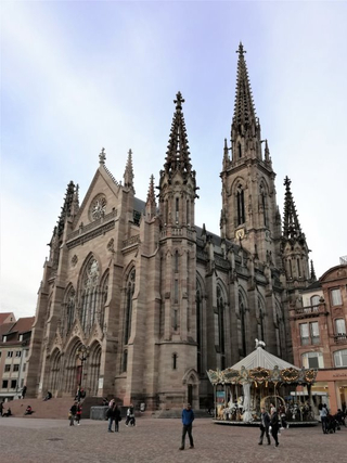 Eine große gotische Kirche mit Doppeltürmen und verzierten Fassaden blickt auf einen Stadtplatz mit festlichem Karussell und Spaziergängern.