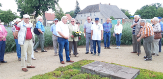 Eine Gruppe älterer Erwachsener steht bei einer Gedenkfeier um einen liegenden Grabstein herum, ein Mann hält Blumen.