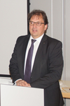 Ein Mann im dunklen Anzug, weißem Hemd und gemusterter Krawatte, mit Brille, steht hinter einem weißen Rednerpult und spricht.