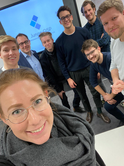Eine lächelnde Frau macht ein Selfie mit sechs lächelnden Männern vor einem Bildschirm mit dem Junge GSP Logo.