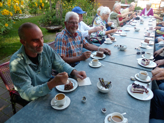 Ältere Menschen genießen Kaffee und Kuchen an einem langen Tisch im Garten, umgeben von gelben Blumen.