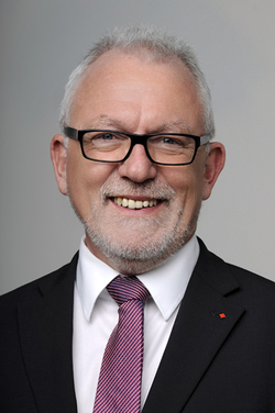 Lächelnder Mann mit grauem Haar, Bart und Brille, trägt Anzug und lila Krawatte.