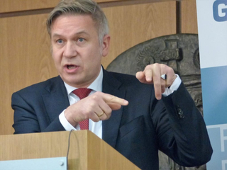 Ein Mann im Anzug spricht am Rednerpult und gestikuliert mit beiden Händen. Er trägt einen markanten Ring.
