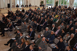Ein vielfältiges Publikum aus Zivilisten und Militärpersonal sitzt in Reihen bei einer formellen Konferenz oder Veranstaltung.