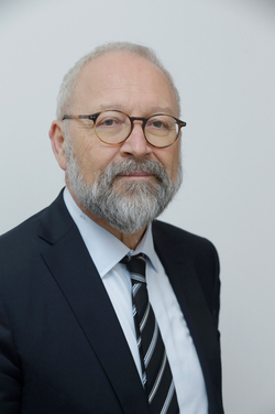 Porträt eines älteren Mannes mit grauem Haar, grauem Bart, Brille, dunklem Anzug, hellblauem Hemd und gestreifter Krawatte.