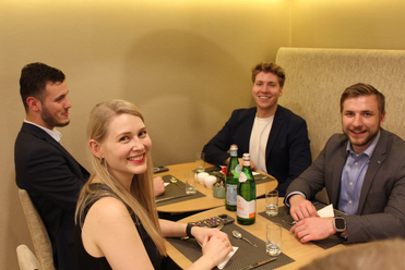 Vier lächelnde Menschen, drei Männer und eine Frau, an einem Restauranttisch mit Getränken.