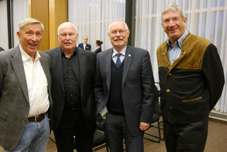 Vier lächelnde ältere Herren, teils in Anzügen, teils in eleganter Freizeitkleidung, stehen nebeneinander in einem Raum.