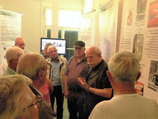 Männer in einer Ausstellung zur deutschen Sicherheitspolitik; ein älterer Mann mit Schnurrbart erklärt etwas, die Gruppe lauscht.