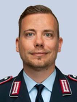Kopfaufnahme eines Mannes in dunkelblauer Uniform mit rot-weißen Rangabzeichen, kurzen braunen Haaren und kurzem Bart.