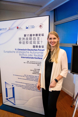 Lachende blonde Frau in weißer Strickjacke vor einem Banner des 11. Chinesisch-Deutschen Forums.