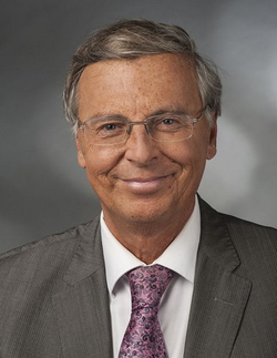 Porträt eines lächelnden älteren Mannes mit grauem Haar und Brille, der einen grauen Anzug und eine rosa Krawatte trägt.