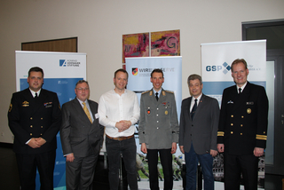 Sechs Männer, teils in Uniform, teils in Anzügen, stehen vor Bannern einer politischen Stiftung und von Reserveverbänden.