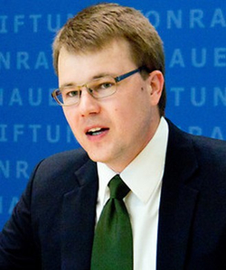 Ein Mann mit Brille und grüner Krawatte spricht vor blauem Hintergrund mit teilweisem Text 'Konrad Adenauer Stiftung'.
