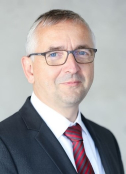 Mann mittleren Alters mit Brille, grauen Haaren und einem dunklen Anzug mit rot-grauer Krawatte, blickt in die Kamera.