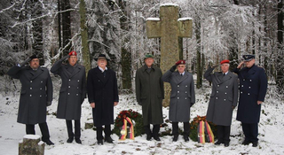 Veteranen in Uniform salutieren an einem verschneiten Waldgedenkort mit Steinkreuz und deutschen Kränzen.