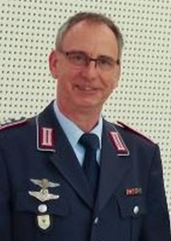 Lächelnder Mann in dunkelblauer Militäruniform mit Fliegerabzeichen, Schulterinsignien und Brille, vor hellem gepunktetem Hintergrund.