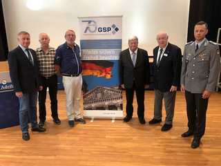Sieben Männer, einer in Militäruniform, stehen vor einem GSP 70-Jahre-Banner mit der Aufschrift 'Frieden sichern, Freiheit bewahren'.