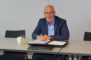 Lächelnder Mann im blauen Anzug mit Brille und Schlüsselband, sitzt am Konferenztisch mit Dokumenten, Laptop und Getränken.