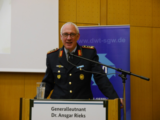 Generalleutnant Dr. Ansgar Rieks in Militäruniform spricht lächelnd hinter einem Rednerpult.