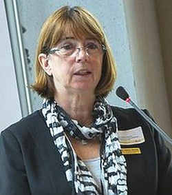Eine Frau mit Brille und gemustertem Schal spricht an einem Mikrofon während einer Präsentation.