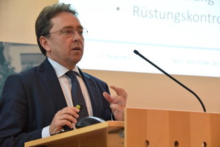 Ein Mann im Anzug und mit Brille spricht an einem Rednerpult und gestikuliert. Im Hintergrund: 'Rüstungskontrolle'.