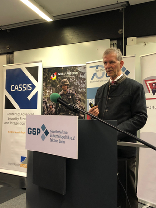 Ein Mann in dunkler Jacke spricht an einem Rednerpult mit Mikrofon. Banner von CASSIS, WIR SIND DIE RESERVE und GSP sind zu sehen.