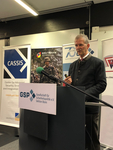 Ein Mann in dunkler Jacke spricht an einem Rednerpult mit Mikrofon. Banner von CASSIS, WIR SIND DIE RESERVE und GSP sind zu sehen.