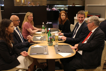 Sechs Personen in Geschäftskleidung, drei Männer und drei Frauen, sitzen an Tischen in einem Restaurant und unterhalten sich.
