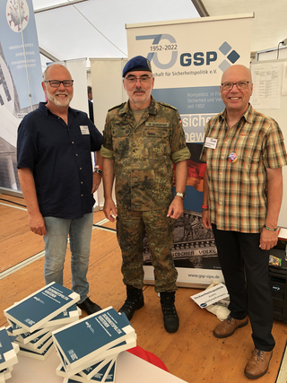 Drei Männer, einer in Uniform, zwei zivil, lächeln an einem GSP-Stand mit Büchern über Sicherheitspolitik.