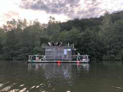 Holz-Hausboot auf einem ruhigen See oder Fluss, mit Menschen, die auf der Dachterrasse entspannen, umgeben von Wald unter bewölktem Himmel.