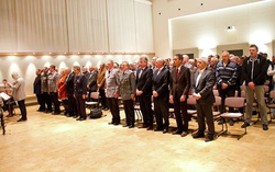 Männer in Anzügen und Uniformen stehen formell in einem Saal, blickend auf Musiker links.